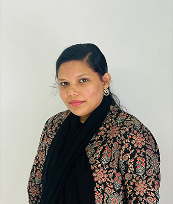 Young professional woman in
									Kerala, dressed in a black and floral-patterned outfit with a matching scarf,
									standing against a plain light background, exuding a calm and confident demeanor.