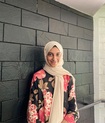 Young professional woman in Kerala, dressed in a cream-colored hijab and a floral-patterned outfit, standing against a dark stone wall, radiating a warm and composed expression.