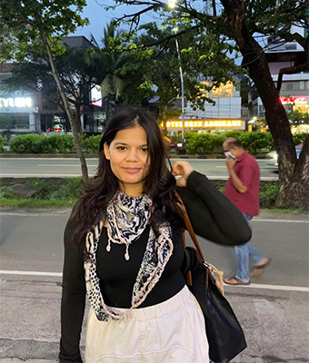 A young woman with long, dark hair and a patterned scarf is standing on a city street at dusk. She wears a black top and a white skirt and gives a soft smile.