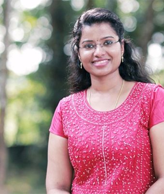 A young woman with glasses and dark, wavy hair is wearing a bright pink top with white beadwork. She is smiling warmly against a soft-focus green outdoor background, giving a professional and pleasant look.