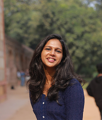 A young woman with long, wavy dark hair and a blue textured top is standing in a sunny outdoor setting near a historical-looking building. She is smiling broadly, conveying a friendly and energetic presence.
