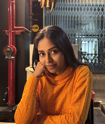 Young professional woman in
									Kerala, dressed in a black and floral-patterned outfit with a matching scarf,
									standing against a plain light background, exuding a calm and confident demeanor.
