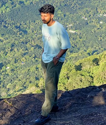 Software developer in Kerala wearing a light blue t-shirt and olive trousers, standing on a rocky cliff edge overlooking lush green hills and valleys.