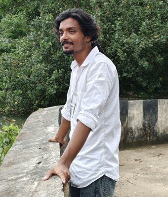 Software developer in Kerala with long tied-back hair, wearing a white shirt, leaning on a canal-side wall with green trees and backwaters behind him.