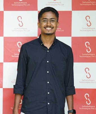Smiling software developer in Kochi, Kerala, wearing glasses, a dark navy shirt and a white mundu, standing in front of the Saiha Software Technologies backdrop.