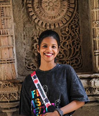 Young creative professional in Kerala, dressed in a casual dark t-shirt with colorful prints, standing before an intricately carved stone backdrop, showcasing a bright and friendly smile.