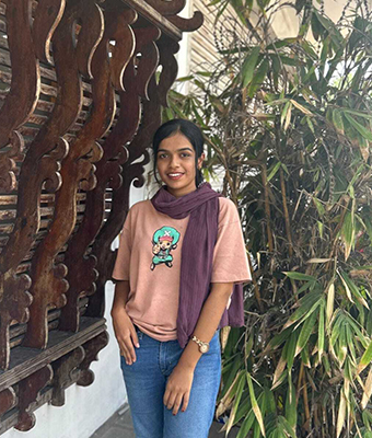 A young woman with long, dark hair is posing outdoors next to a wooden, carved screen and some bamboo. She wears a casual pink t-shirt with a graphic print, blue jeans, and a long maroon scarf