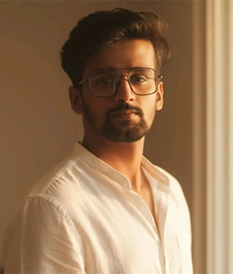 A young man with a beard and dark, modern glasses is wearing a crisp white collared shirt. He is posed indoors with warm, natural light illuminating his face, giving a serious and contemplative look.