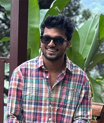 A young man with dark sunglasses and a beard is wearing a colorful plaid shirt. He is seated outdoors in a lush, green setting and is smiling brightly, radiating a casual and joyful expression.