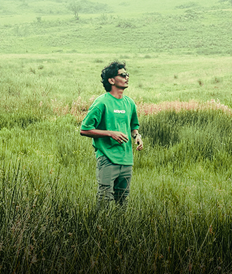 A young man with curly hair and sunglasses is standing in a vast, overgrown green field. He is dressed in a monochromatic green outfit (t-shirt and pants), looking up and away, suggesting a pensive and adventurous mood.