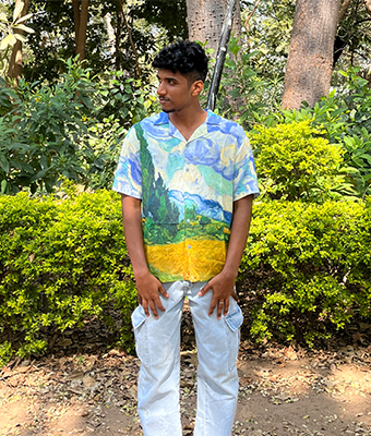 A young man with curly hair is standing outdoors in a park-like setting with green bushes. He is wearing a distinctive short-sleeve shirt featuring an artistic, painted landscape design and light-colored cargo pants, looking off to the side.