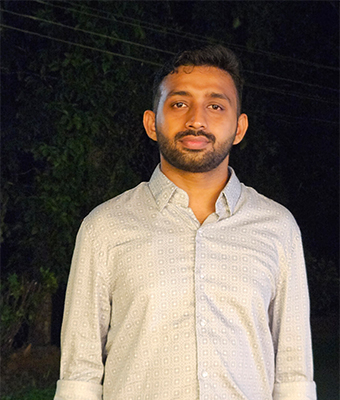 A young man with a short beard is wearing a light gray shirt with a subtle geometric pattern. He is standing outdoors at night with dark foliage in the background, looking directly at the camera with a neutral expression.